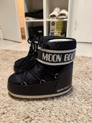 Svarta Moon Boot med vit logga - Säljer ett par klassiska svarta Moon Boot med vit logga och detaljer. Boots i nylon och syntetmaterial med snörning framtill och hög sula. Perfekta för kalla dagar och riktigt snygga till vinterlooken.