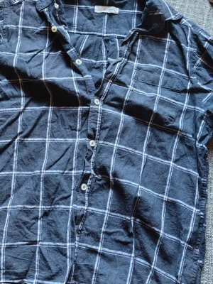 Rutig svart skjorta från S.C.W - Säljer en svart skjorta från S.C.W med vitt rutmönster. Skjortan har klassisk krage, bröstficka och knappar framtill. Tillverkad i ett lätt och luftigt material, perfekt för en avslappnad stil. Säljer då jag vuxit ur den andvända fåtal gånger 