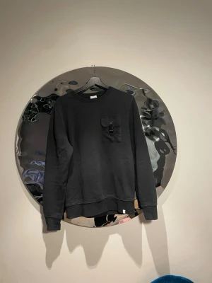 Svart sweatshirt med ficka och spänne - Cool svart sweatshirt med rund hals och lång ärm. Bröstficka med lock och ett snyggt spänne som detalj. Perfekt för dig som gillar minimalistisk stil med en twist. Mjuk bomullskänsla och ribbade muddar vid ärmslut och nederkant.