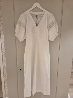 Vit långklänning från Soyaconcept - Långklänning i vitt från Soyaconcept med v-ringning och puffärmar. Klänningen har fina spetsdetaljer på ärmarna och är tillverkad polyester och bomull. Perfekt för sommarens alla tillfällen.