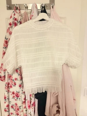 Vit croppad blus med volang - Säljer en söt vit blus med croppad passform och volangkant nedtill. Blusen har korta ärmar och ett skirt, strukturerat tyg som ger en luftig känsla. Perfekt till höga jeans eller kjol för en trendig look.