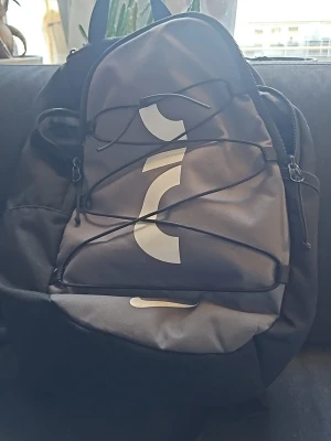 Svart Nike Air ryggsäck - Snygg svart ryggsäck från Nike Air med vit logga och detaljer. Ryggsäcken har ett stort huvudfack, justerbara axelband och elastisk snörning på framsidan. Tillverkad i slitstarkt material, perfekt för skola eller träning.