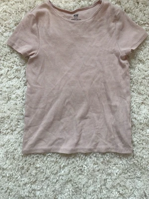 Ljusrosa ribbad t-shirt från H&M - En enkel och stilren ljusrosa t-shirt från H&M med ribbad struktur. T-shirten har rund halsringning och korta ärmar. Tillverkad i mjukt och stretchigt material som sitter skönt på kroppen. Perfekt basplagg för alla tillfällen.