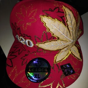 Röd keps med broderade löv XL - Säljer en röd keps från Leader i storlek XL med stora broderade löv i guld, beige och svart. Kepsen har även siffran 420 broderad framtill och detaljer i metallictråd. Perfekt för dig som gillar statement-plagg och unik stil.