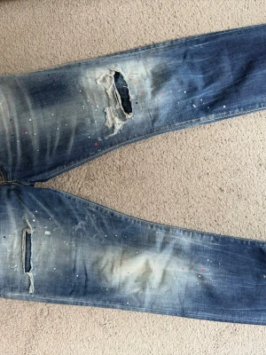 Dsquared2 jeans  - Säljer ett par blå jeans med slitningar och stora hål på knäna. Jeansen har en tvättad look med ljusa partier och små färgstänk i vitt och rött över hela benen. Rak passform och klassiska bakfickor. Perfekt för dig som gillar en edgy och trendig stil.