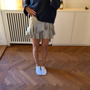 Glittrig volangkjol i silver - Supersnygg och gullig kort glittrig kjol i silver med volangdetaljer. Kjolen har en flowy passform och är perfekt för dig som vill sticka ut. Materialet känns lätt och skimrande, vilket ger en extra festlig känsla.