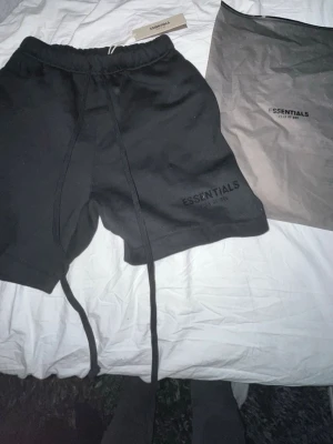 Svarta shorts från Essentials -  Helt oörda med tags kvar. Svarta shorts från Essentials med långa snören i midjan och diskret logga på vänster ben. Mjukt material och relaxed fit, perfekta för chill dagar. Elastisk midja för extra komfort.