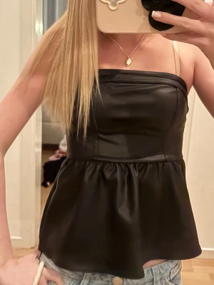 Svart satinlinne med tunna axelband - Snyggt svart linne i satin med tunna axelband och rak urringning. Toppen har en figurnära överdel och ett lätt utställd nederdel som ger en cool siluett. Perfekt att styla med jeans eller kjol för en trendig look.