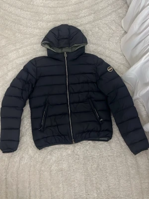 Svart pufferjacka från Colmar - Snygg svart pufferjacka från Colmar med huva och dragkedja framtill. Jackan har två fickor med dragkedja och en rund Colmar-logga på ärmen. Perfekt för kalla dagar och riktigt stilren look.