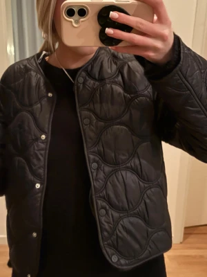 Svart quiltad cardiganjacka - Stilren svart quiltad cardiganjacka med rund halsringning och tryckknappar framtill. Jackan har ett vågigt mönster i sömmarna och är lätt vadderad för extra komfort. Perfekt för lager på lager och ger en trendig look.