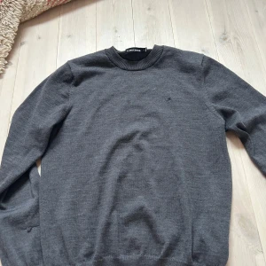 Mörkgrå stickad tröja J.Lindeberg - Mörkgrå stickad tröja från J.Lindeberg med rund halsringning och diskret logga broderad på bröstet. Tröjan har en klassisk passform och ribbade muddar vid ärmslut och nederkant. Perfekt för lager på lager och stilren look.