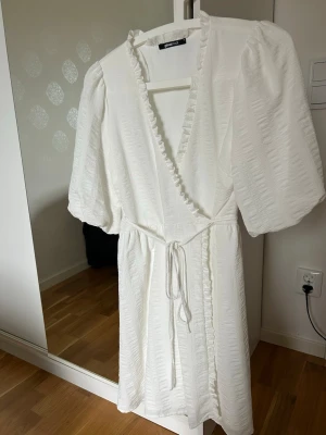 Vit omlottklänning från Gina Tricot - Supersöt vit omlottklänning från Gina Tricot med puffärmar, volangkant längs v-ringningen och knytband i midjan. Klänningen har en lätt struktur i tyget och känns luftig och somrig. Perfekt för varma dagar och stilrena outfits.