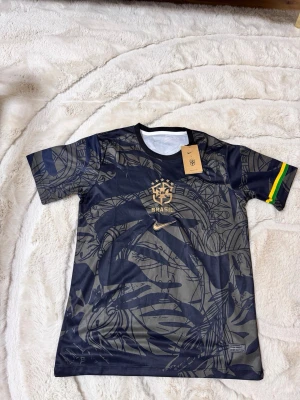 Brasil Nike matchtröja svart/guld - Snygg svart matchtröja från Nike med Brasil-logga och guldiga detaljer. Tröjan har ett abstrakt mönster, rund hals och korta ärmar. På ena ärmen finns en rand i grönt, gult och blått. Perfekt för fotboll eller streetwear. 