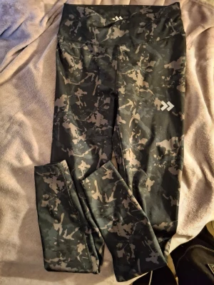 Svarta camo leggings från 157 - Säljer ett par svarta leggings från 157 med grått camomönster. De har hög midja och är tillverkade i ett stretchigt material som sitter skönt på kroppen. Perfekta för träning eller chill. Snygg logga på benet och insidan av linningen.