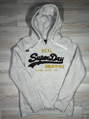 Ljusgrå hoodie från Superdry - Snygg ljusgrå hoodie från Superdry med stor logga i svart och gult framtill🤩Hoodien är i fint skick och passar Xs-S🙌🏻 Hör av er vid funderingar samt snabbaffär😇