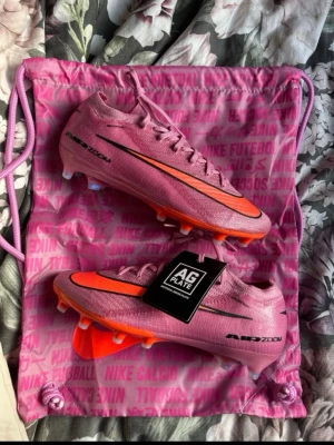 zm vapor 16 elite ag-pro nike fotbollsskor - Säljer ett par Nike Mercurial fotbollsskor i rosa med orange och svarta detaljer. Skorna har AG Plate-sula för konstgräs och är tillverkade i ett lätt syntetmaterial. Snygg design med Air Zoom-teknologi och klassisk Nike-logga på sidan. Perfekta för dig som vill sticka ut på planen. Ordinarie pris är 3099kr. Aldrig använda och helt nya