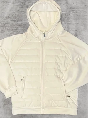 Vit Cardigan - Snygg vit cardigan med quiltade paneler framtill och baktill. Tröjan har hel dragkedja, två fickor med dragkedja och en skön huva. Perfekt för kyliga dagar och enkel att matcha med det mesta. Långärmad och med ribbade muddar vid ärmslut och nederkant. Passar perfekt till dig som är över 170cm lång