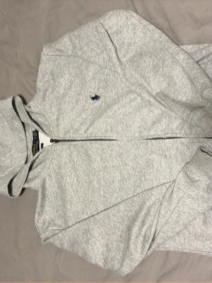 Grå zip hoodie från Polo Ralph Lauren - Snygg gråmelerad hoodie från Polo Ralph Lauren med dragkedja framtill och klassisk blå logga på bröstet. Hoodien har huva, känguruficka och ribbade muddar vid ärmar och nederkant. Perfekt för en chill och avslappnad stil. Priset kan diskuteras!!