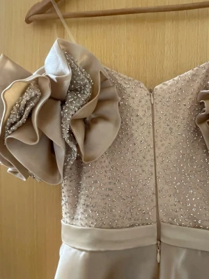 Champagnefärgad långklänning med volang - Elegant långklänning i champagnefärg med glittriga pärldetaljer på överdelen och stora volangärmar. Klänningen har axelbandslös design och är tillverkad i ett glansigt material som ger en lyxig känsla. Perfekt för bal eller festliga tillfällen.