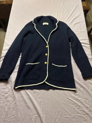 Marinblå kavaj med vita detaljer - Stilren marinblå kavaj från GANT med vita kontrastsömmar längs krage, fickor och knappslå. Tre guldknappar framtill och två framfickor. Mjuk, stickad känsla och klassisk passform som ger en avslappnad men ändå uppklädd look.