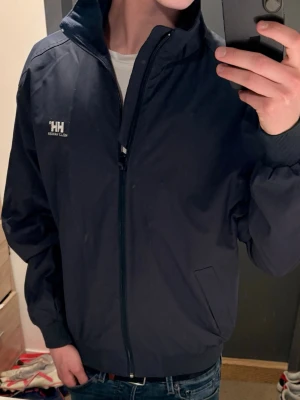 Mörkblå vindjacka Helly Hansen - Snygg mörkblå vindjacka från Helly Hansen med diskret HH-logga på bröstet. Storlek M 