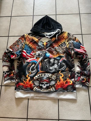 Färgstark hoodie med motorcykeltryck storlek L - Säljer en riktigt fet hoodie med stort tryck av en örn, motorcykel, amerikanska flaggor och eldflammor. Huvan är svart med vita snören och resten av hoodien är täckt av detaljerade motiv i rött, blått, orange och vitt. Perfekt för dig som gillar streetwear och statement-plagg.