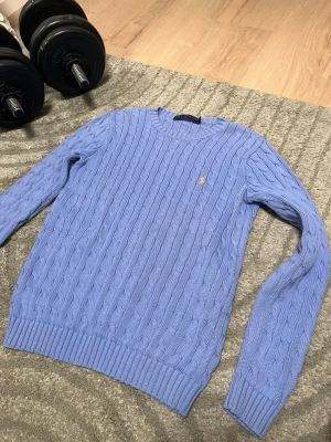 Ljusblå kabelstickad tröja Ralph Lauren - Snygg ljusblå kabelstickad tröja från Ralph Lauren Sport med rund halsringning och diskret broderad logga på bröstet. Tröjan har långa ärmar och ribbade muddar vid ärmslut och nederkant. Perfekt för dig som gillar klassisk preppy stil.