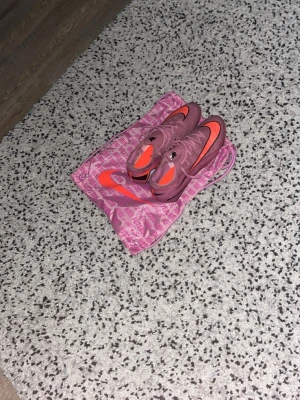 Nike rosa Mercurial fotbollsskor - Säljer ett par rosa Nike Mercurial fotbollsskor med orange detaljer och stora vita M-loggor på hälen. Skorna har snörning och är designade för bra grepp på planen. Medföljer en matchande rosa påse med Nike-logga. Perfekta för dig som vill sticka ut på planen.