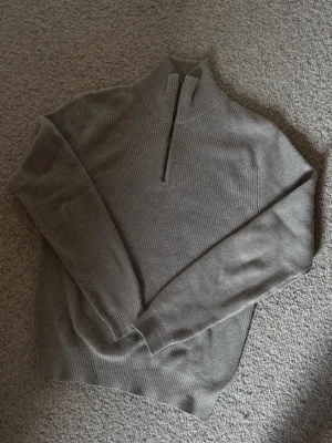 Beige stickad halvzip från Dobber - Säljer en beige stickad tröja från Dobber med halvzip och hög krage. Tröjan har ribbstickad struktur och långa ärmar. Perfekt för lager på lager och enkel att matcha med olika outfits.