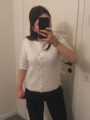 Vit kofta med pärlknappar - Söt vit kofta med korta ärmar och pärlknappar framtill. Koftan har en mjuk och fluffig känsla, rund halsringning och rak passform. Perfekt att styla till både jeans och kjol för en clean look.