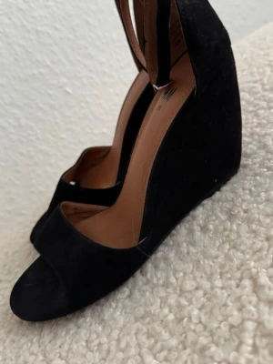 Svarta kilklackade pumps i mocka - Säljer ett par svarta pumps med kilklack och öppen tå. Skorna har en smal rem med spänne runt hälen och är gjorda i mockaimitation. Insidan är beige och sulan är lätt vadderad för extra komfort. Perfekta för dig som gillar stilrena och klassiska skor. Använd 2 gånger. Kilklack 8cm.