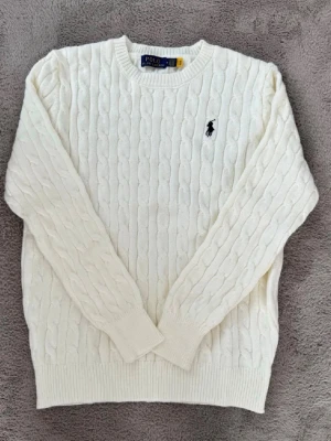 Vit kabelstickad tröja Polo Ralph Lauren - Kabelstickad vit tröja från Polo Ralph Lauren med klassisk rund halsringning och svart broderad logga på bröstet. Tröjan har ribbade muddar vid ärmslut och nederkant. Perfekt för en stilren och tidlös look.
