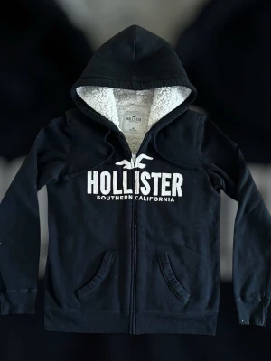 Hollister hoodie - Svart hoodie från Hollister med vit text och logga på bröstet. Mjuk teddyfoder på insidan av huvan och dragkedja framtill. Två fickor fram och ribbade muddar vid ärmar och nederkant. Perfekt för chill dagar.