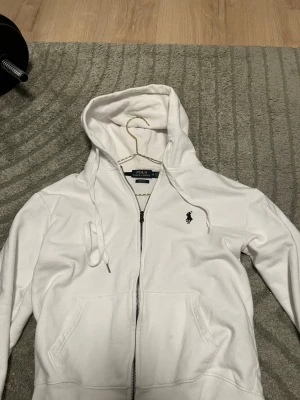 Vit hoodie från Polo Ralph Lauren - Säljer en vit hoodie från Polo Ralph Lauren med dragkedja framtill och klassisk svart logga broderad på bröstet. Hoodien har huva med dragsko och två stora fickor framtill. Tillverkad i mjuk bomull för skön känsla.