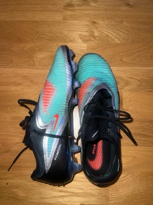 Nike Phantom GT2 turkosa fotbollsskor - Säljer ett par Nike Phantom GT2 fotbollsskor i turkos med svarta och röda detaljer. Skorna har snörning, syntetiskt material och dobbar för spel på gräs. Snygg design med röda Nike-loggor och svart häl. Perfekta för dig som vill sticka ut på planen.