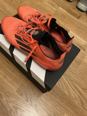 Röda Adidas fotbollsskor med dobbar - Säljer ett par röda Adidas fotbollsskor med svarta detaljer och klassiska tre ränder på sidan. Skorna har snörning, låg vikt och en strumpliknande krage för extra stöd. Ovandelen är i syntetmaterial och sulan har fasta dobbar för spel på gräs.