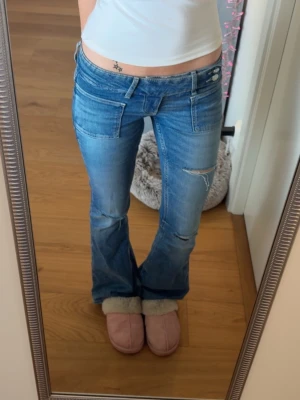 Blå low waist bootcut jeans - Säljer ett par blå low waist jeans med bootcut-modell och slitna detaljer på benen. Jeansen har dubbla knappar i midjan och stora framfickor.