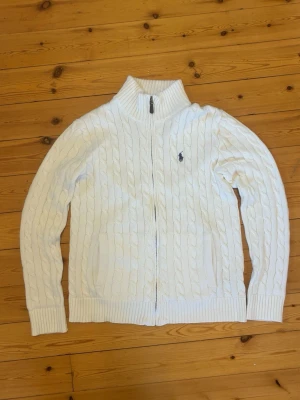 Vit kabelstickad ziptröja Polo Ralph Lauren - Kabelstickad vit tröja med dragkedja från Polo Ralph Lauren. Tröjan har hög krage, ribbade muddar och det klassiska broderade logotypen på bröstet. Perfekt för en clean och stilren look. Materialet känns mjukt och håller värmen.
