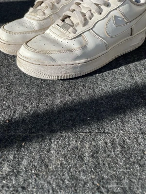 Vita Nike Air Force 1 sneakers - Klassiska vita Nike Air Force 1 sneakers i läder med perforerad tå, vit sula och matchande vita skosnören. Swoosh-logga på sidan och metallbricka med 'AF1' på snörningen. Perfekt för dig som gillar en tidlös och clean stil.