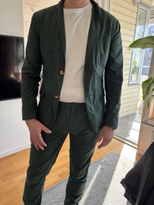 Mörkgrön kostym med kavaj - Snygg mörkgrön kostym med matchande kavaj och byxor. Kavajen har två bruna knappar och klassisk krage. Tillverkad i ett lätt material som ger en avslappnad men stilren look. Perfekt för dig som vill sticka ut med färg men ändå hålla det elegant.