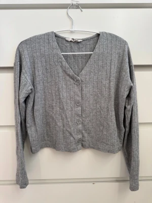 Grå ribbad kofta med knappar - H&M - Mysig grå kofta med ribbad struktur och v-ringning. Koftan har långa ärmar och stängs med fyra stora knappar framtill. Perfekt att slänga över en t-shirt för en chill look. Materialet känns mjukt och stretchigt.