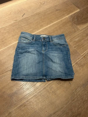 Blå jeanskjol från Zara Woman - Säljer en klassisk blå jeanskjol från Zara Woman med fram och bak fickor och knappgylf. Kjolen har en kort passform och är tillverkad i slitstarkt denimtyg. Snygga detaljer med broderade bakfickor och bälteshällor. Perfekt till vardagslooken!