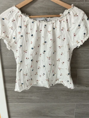 Vit blommig offshoulder topp - Söt vit offshoulder topp med små blommor i blått och rosa. Puffiga korta ärmar och smockad resår upptill ger en romantisk vibe. Perfekt till sommaren💖