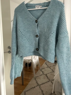 En stickad kofta från Nakd i ljusblå färg med mörkare knappar  - En ljusblå kofta från Nakd som aldrig är använd. Mörkare knappar. Den har en croppad passform.