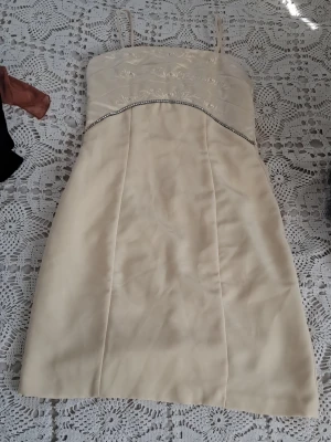 Champagnefärgad kort klänning med pärldetaljer - Elegant kort klänning i champagnefärg med tunna axelband. Överdelen har broderade blommor och en rad med pärlor under bysten som ger extra lyxig känsla. Klänningen har en figurnära passform och är tillverkad i ett slätt, glansigt material.