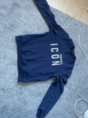 Mörkblå sweatshirt Dsquared2 ICON - Mörkblå sweatshirt från Dsquared2 med stor vit text 'ICON' på bröstet och märkesnamnet tryckt i mindre stil. Tröjan har rund halsringning, långa ärmar och ribbade muddar. Tillverkad i mjukt bomullsmaterial för en skön och avslappnad look. Varan är i strl 16 years men passar storlek S-M.