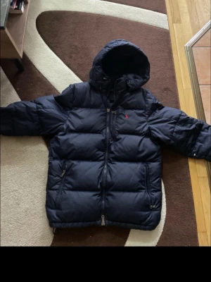 Mörkblå dunjacka från Polo Ralph Lauren -  Köpt från Zalando förra året så den är 100% Äkta, Snygg mörkblå dunjacka från Polo Ralph Lauren med huva och dragkedja framtill. Jackan har två sidofickor med dragkedja och den klassiska röda loggan broderad på bröstet. Perfekt för kalla dagar och riktigt stilren.