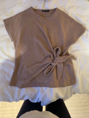 Beige topp med knyt från Zara - Säljer en beige topp från Zara med korta ärmar och snygg knytning i sidan. Toppen har en rund halsringning och är gjord i ett mjukt stickat material som känns skönt mot huden. Perfekt för dig som gillar stilrena och enkla plagg med en twist.