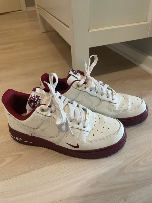 Nike Air Force 1 vita och vinröda - Säljer ett par Nike Air Force 1 sneakers i vitt läder med vinröda detaljer och sula. Skorna har klassisk låg profil, perforerad tå och Nike-logga på sidan. Insidan är fodrad i vinrött tyg och tungan har en matchande logga. Perfekta för dig som gillar stilrena sneakers med färg.