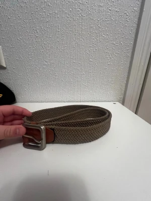 Brunt flätat bälte med skinn-detaljer - Snyggt brunt flätat bälte med detaljer i brunt skinn och en silvrig metallspänne. Bältet har en klassisk look och passar perfekt till jeans eller chinos. Längd 90 cm enligt märkning på läderdetaljen.